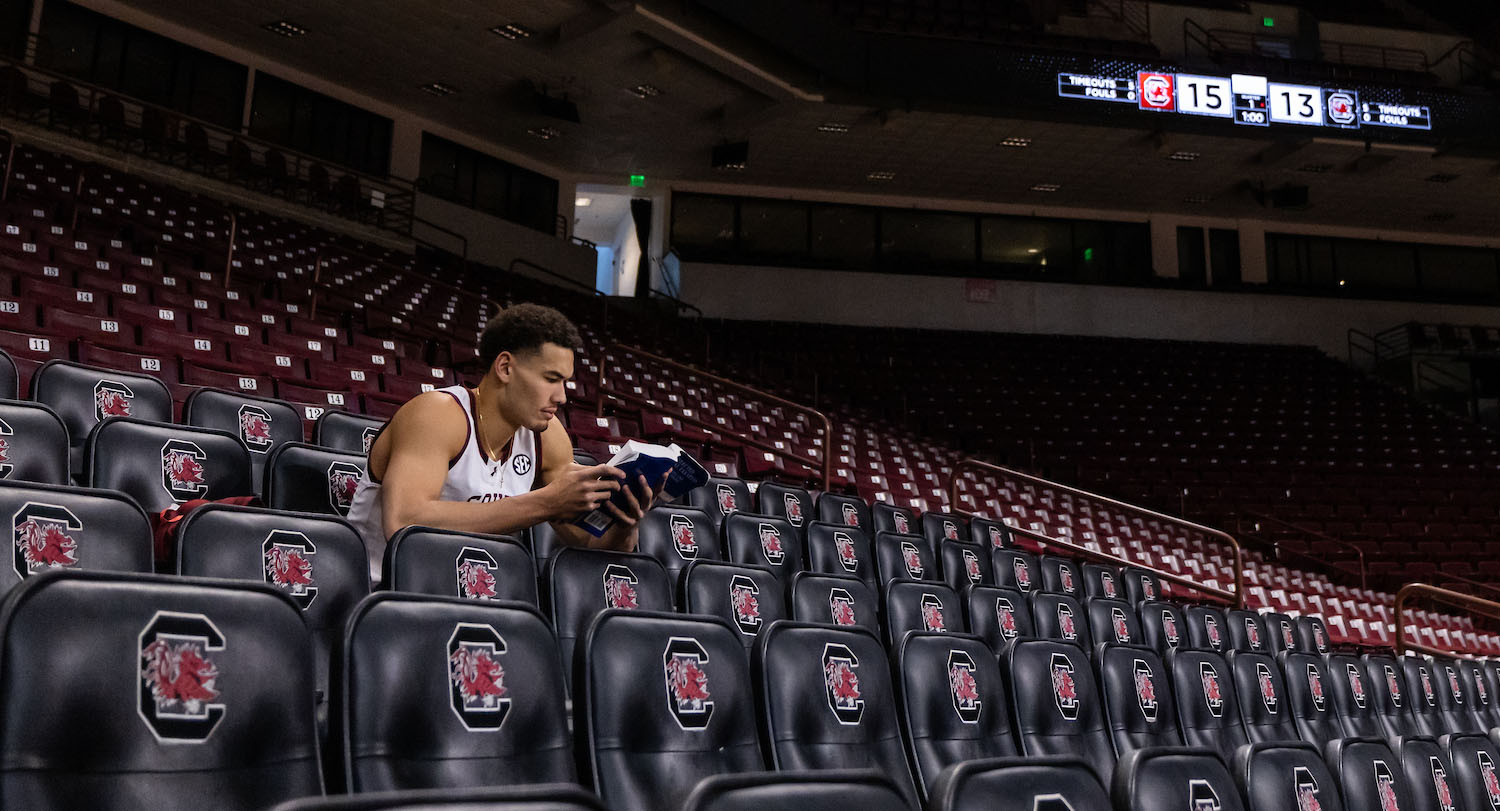 Man in white jersey with garnet South Carolina lettering reads blue textbook in empty basketball arena