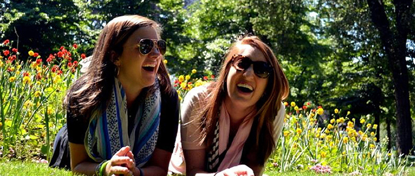 Two female students lay on the grass in front of a bed of flowers on a sunny day, laughing and wearing sunglasses