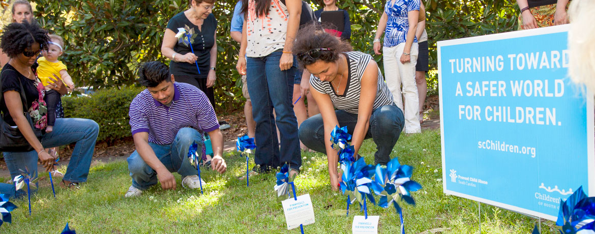 A group of people, including men and women and one woman holding a baby, place blue-and-silver pinwheels in a grassy area beside a sign that reads "Turning toward a safer world for children. scChildren.org"