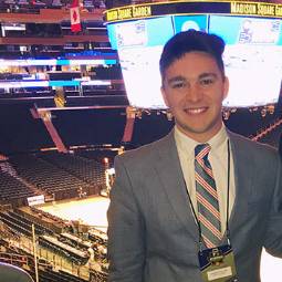 Male student in suit and tie with ID badge on a lanyard, standing in Madison Square Garden arena with Jumbotron in background