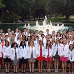 Group of 50 or more students in white lab coats posing for a group shot outdoors in front of a fountain