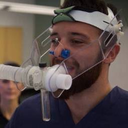 Male student in dark-colored scrubs demonstrates the use of a breathing device on his head and face