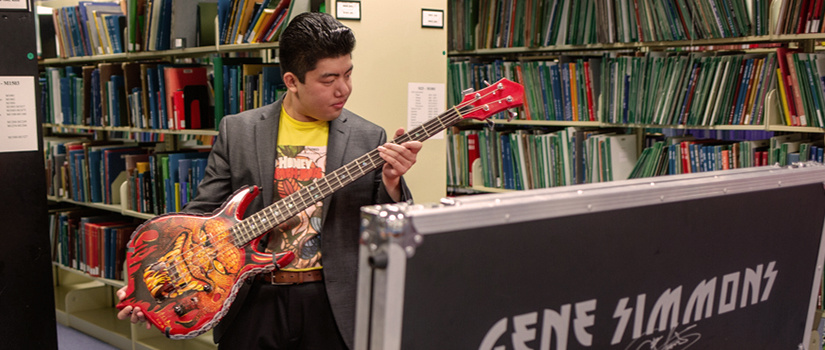 A graduate student in professional clothing holds an epic red guitar in front of a case labeled Gene Simmons and signed by Gene himself.