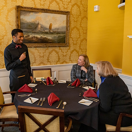 A student working at McCutchen House speaks with two customers who are seated at a table.