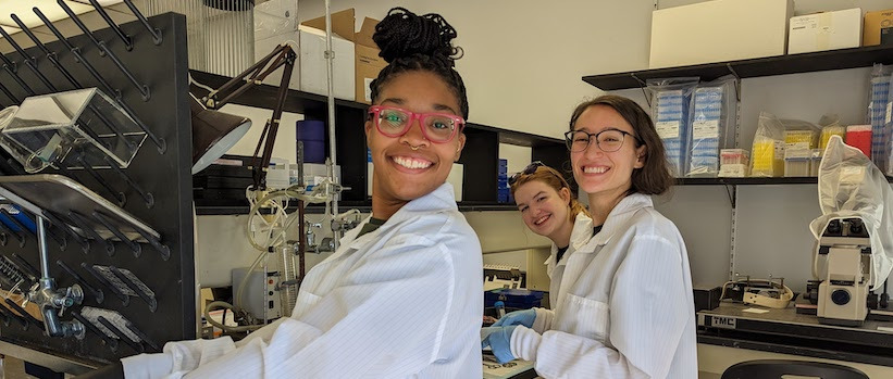 A group of researchers working in the lab smiling for the camera in front of their research equipment