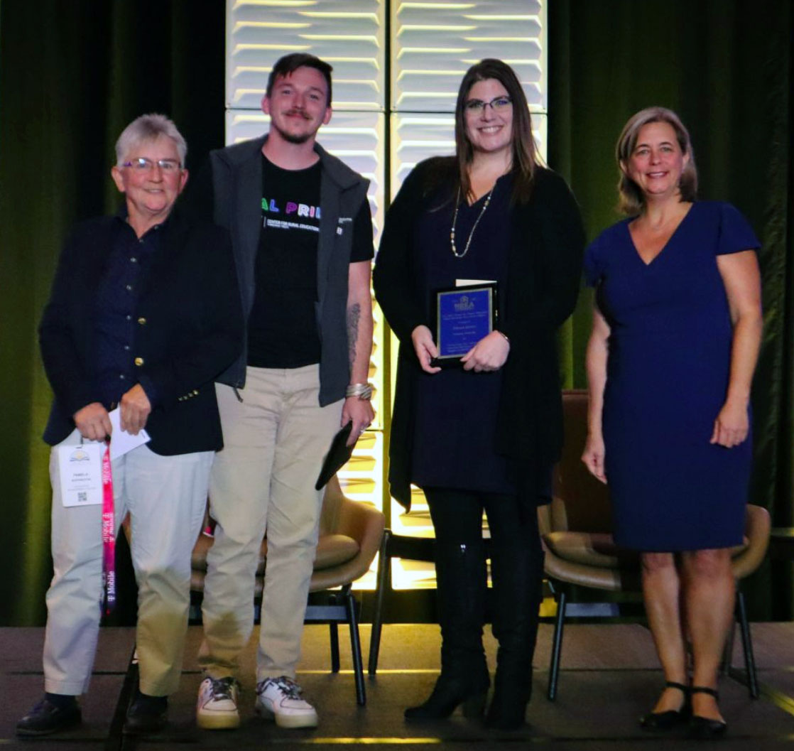 Several smiling people on stage, Devan Jones is second from right, holding an award.