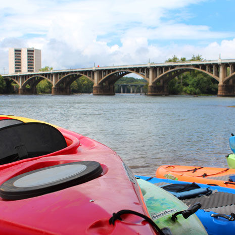 Red, orange, blue, and green kayak floats are pictured on the edge of a river with a bridge and skyscraper building in the background. 