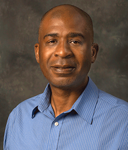 Philip is pictured from the shoulders up wearing a blue collared shirt. He has dark skin and is smiling. 