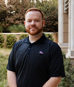 Casey is pictured from the waist up wearing a black Carolina polo. He has red hair, fair skin, and is smiling at the camera. 