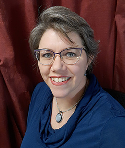 Carrie is pictured from the shoulders up wearing eyeglasses and a blue shirt. She has hair skin and is smiling. 