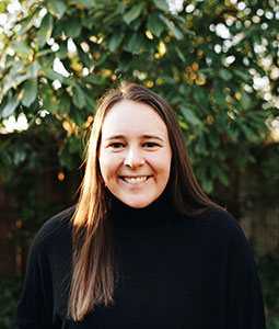 Gray is pictured in front of a tree with green foliage. She is wearing a black turtle neck, has pale skin and long brown hair and is smiling. 