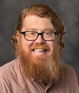 Mike is pictured from the shoulders up. He has red hair and a red beard and is wearing eye glasses. He is smiling. 