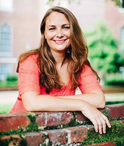 Ali is pictured leaning against a brick wall. She has pale skin, long brown hair and is smiling. 