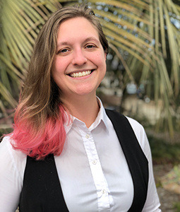Anna is wearing a white shirt and black vest. She has shoulder length blonde hair with pink tips and she is smiling. 