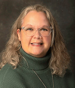 Tina is pictured fromt he shoulders up wearing a green turtleneck sweater and eyeglasses. She has wavy blonde hair, fair skin, and is smiling. 