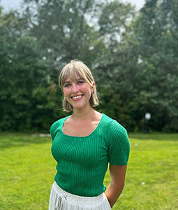 Corinne is pictured standing in front of green trees wearing a green shirt and white skirt. She has fair skin, short blonde hair and is smiling. 