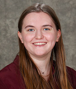 Hannah is pictured from the shoulders up wearing a garnet sweater. She has fair skin, long brown hair and is smiling. 