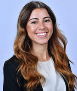 Destinee is pictured from the waist up in front of a purple background. She is wearing a whit shirt and black jacket. She has long brown hair and is smiling. 