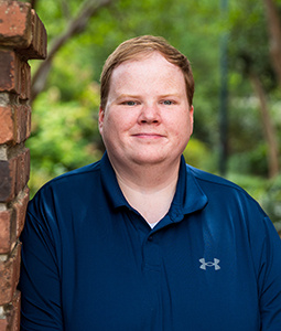 Andrew is pictured from the shoulders up wearing a blue shirt. He has blonde hair and fair skin. 