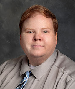 Andrew is pictured from the shoulders up. He is wearing a gray collared shirt with a blue striped tie. 