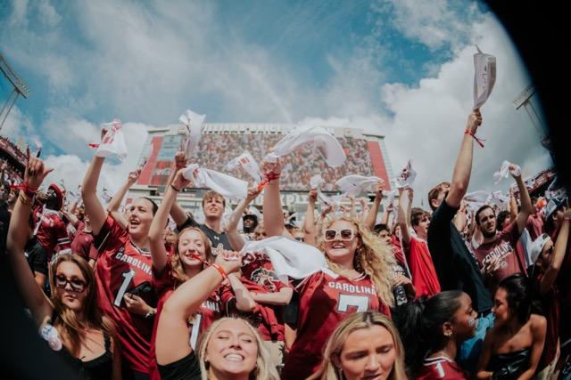Fans wearing Gamecock gear cheer in a crowded stadium, waving white towels.