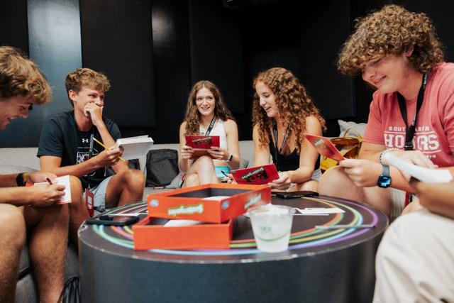 A group of college students sit around a table laughing and playing a card game.