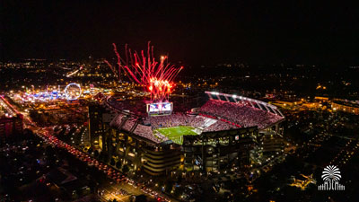 An aerial view of Williams-Brice Stadium during a night game with fireworks shooting from the scoreboard and the state fair in the background with tree and gates mark with tree and gates mark.