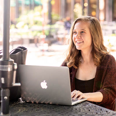 A student looking up and smiling over her laptop at an outdoor table outside of Russell House.