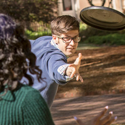 A student throwing a frisbee to another student in the foreground outside.