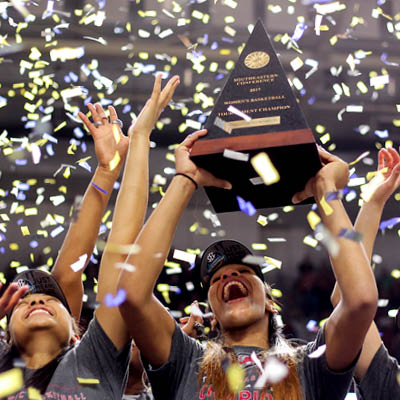 The women's basketball team under confetti holding up a trophy.