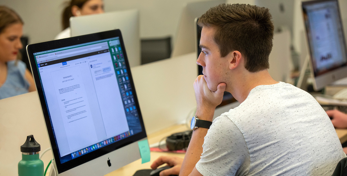 With a hand on his chin, a student gazes at a desktop computer displaying a document that contains text.