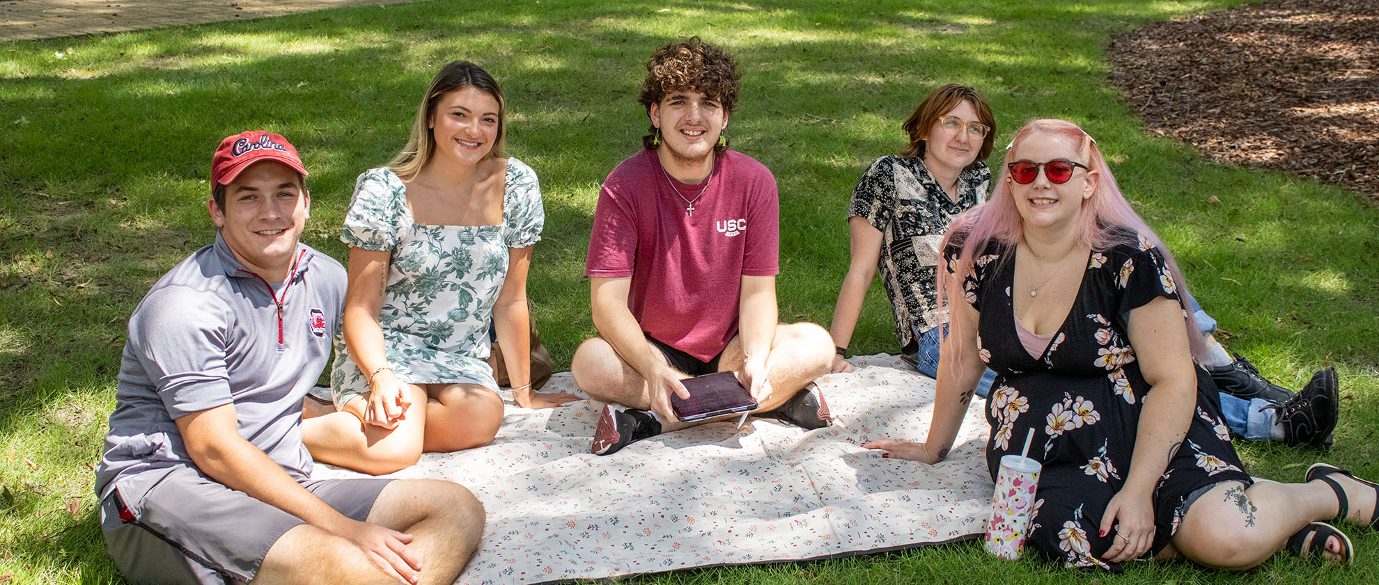 Students lounge on a picnic blanket on the horseshoe, with the student in the middle holding their tablet.