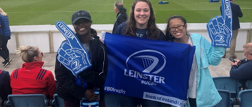 Three students hold up foam fingers and flags at a rugby match