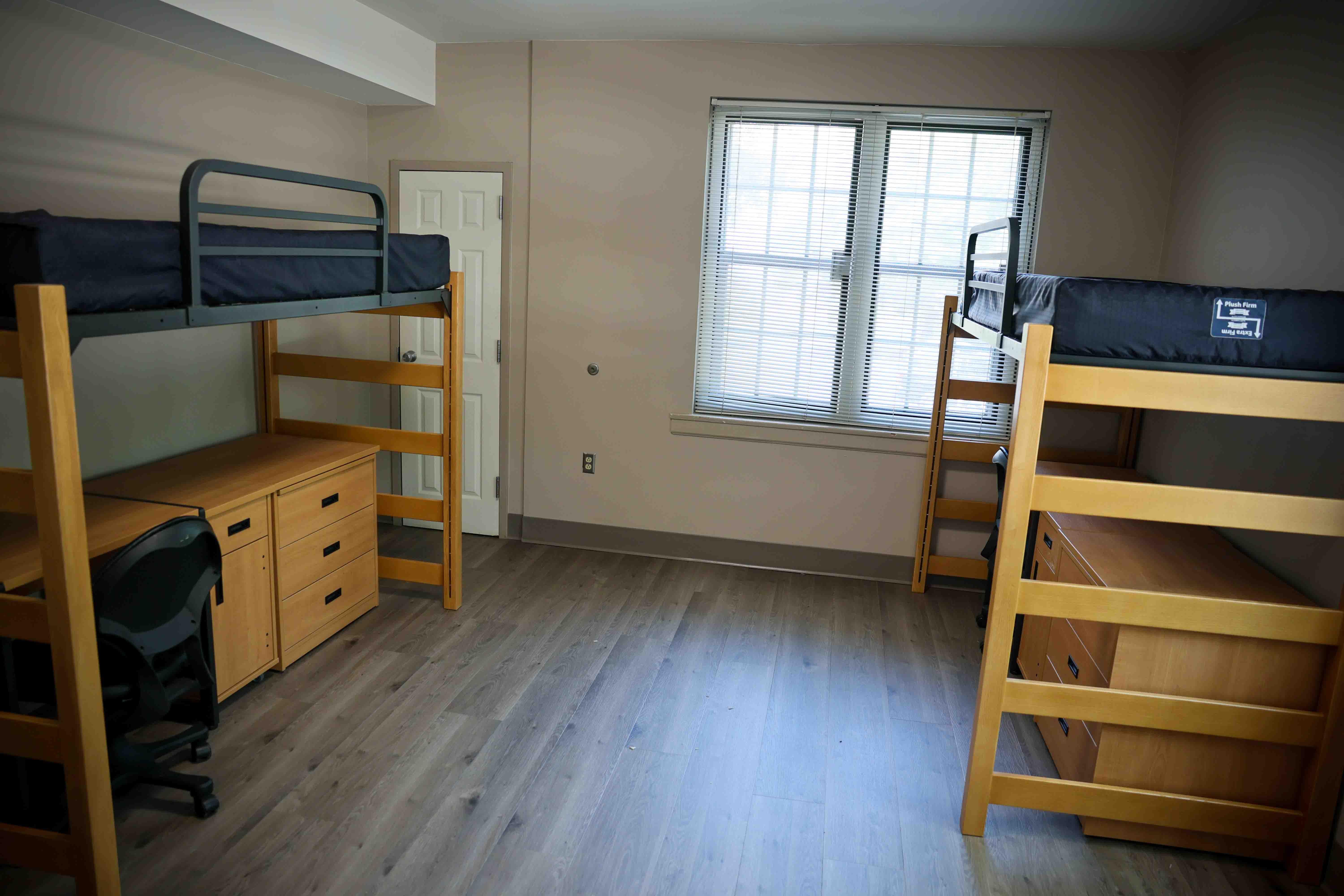 A spacious double-occupancy residence hall room featuring lofted beds, study desks, dressers, and abundant open floor space.