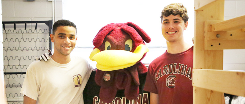two guys posing for a photo with Cocky in their residence hall room
