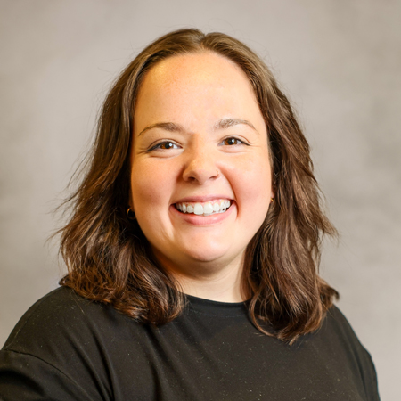 Professional headshot of a smiling person with shoulder-length brown hair wearing a black crew-neck shirt. The person is facing the camera against a soft gray studio background with warm, even lighting, creating a friendly, approachable, and professional appearance.