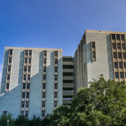 A front view of Bates House, a building with blue/grey bricks and windows lining the building in columns, with green trees in the foreground.