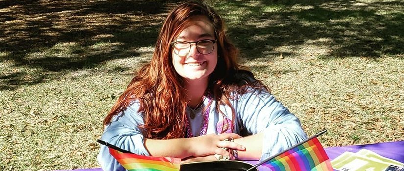 A girl is sitting in front of a table in front of pride flags.