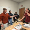 A group of students gathers around a table indoors, talking and participating in an interactive activity.