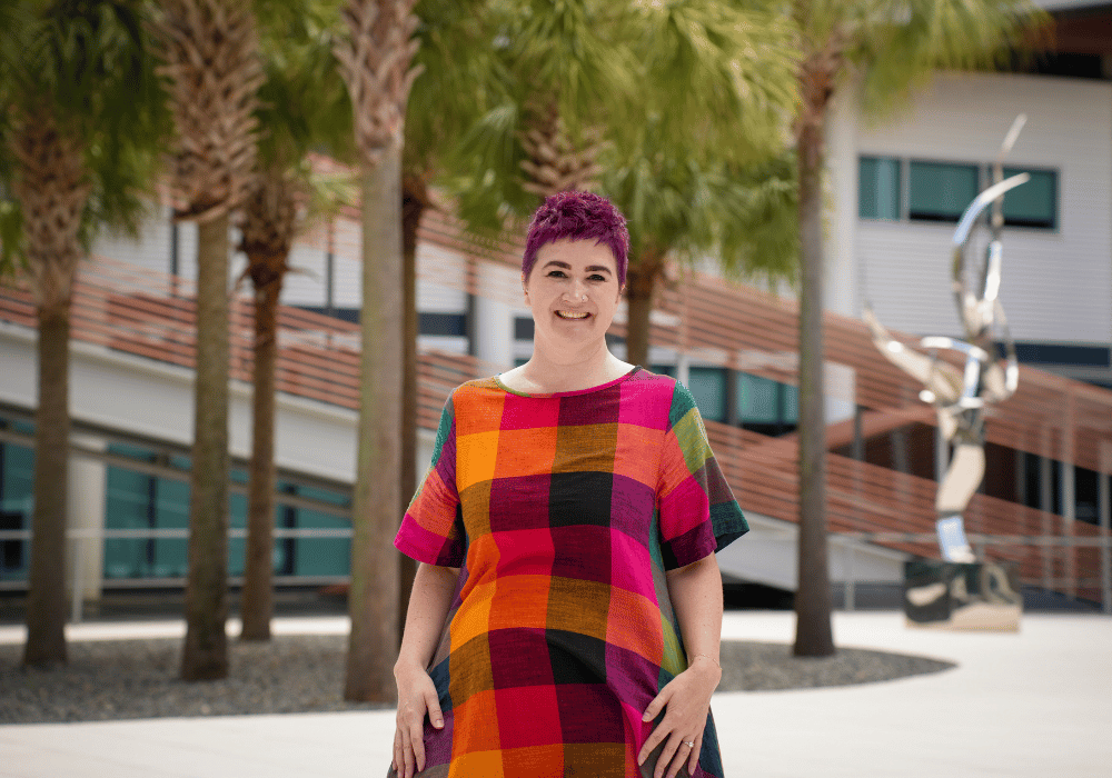 A smiling person with short purple hair stands outdoors on a university campus, wearing a colorful multicolored plaid dress. Palm trees and a modern campus building are visible in the background.