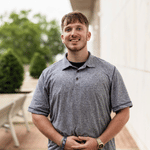 Young man in a gray polo shirt standing outdoors and smiling at the camera.