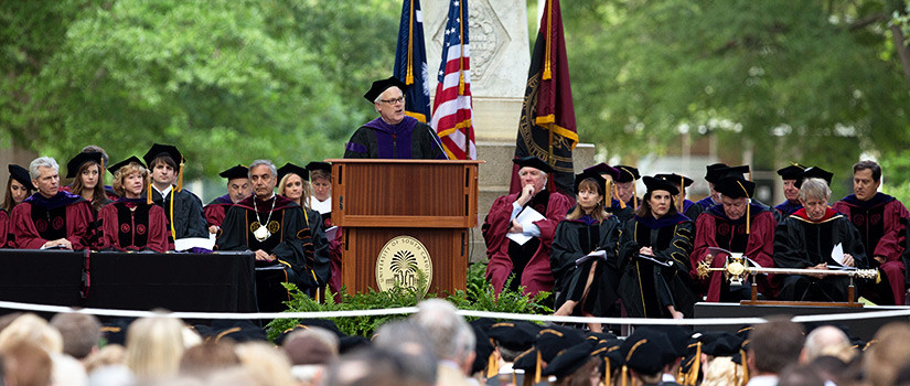 guest speaker standing at podium on stage with faculty on the Horseshoe in front of the Maxcy Monument during law school ceremony