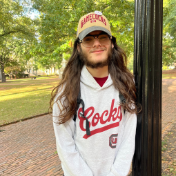 USC undergraduate student researcher Ethan Irizarry poses next to a light post on the historic University of South Carolina Horseshoe. He wears a Gamecocks cap and hooded sweatshirt as he smiles at the camera.