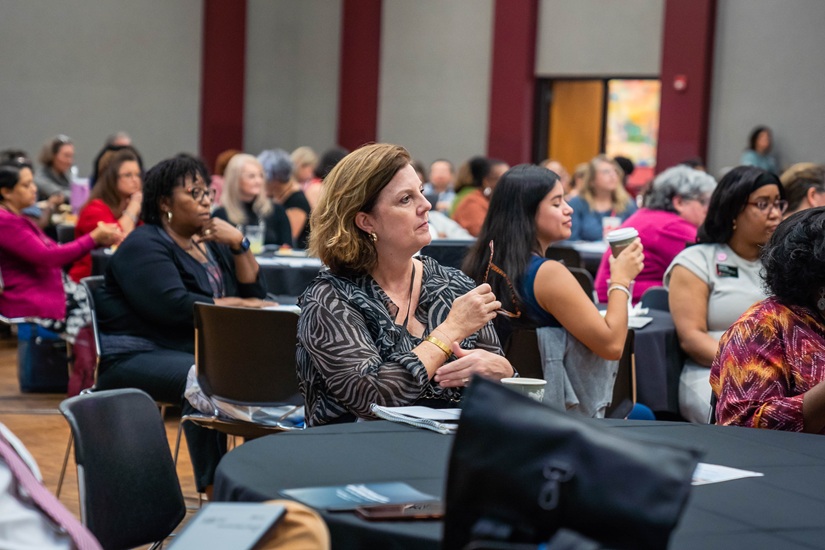 USC research administrator Janie Godbold takes in the conference welcome address while sitting at a round table draped in a black tablecloth, with scores of her fellow attendees visible in the background.. 