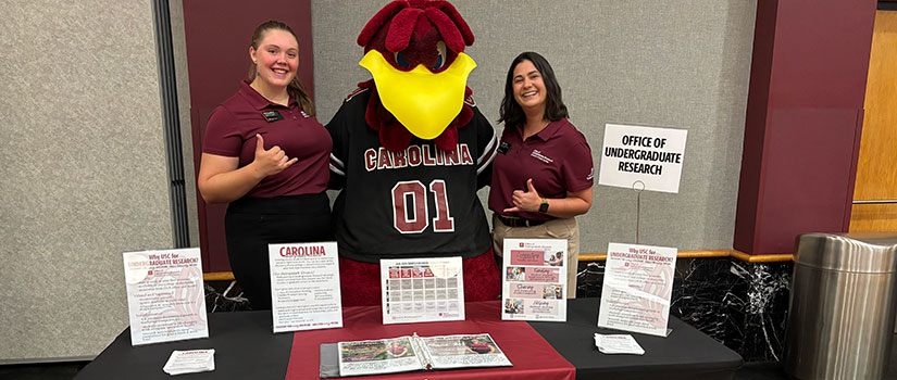 Two Undergraduate Research Ambassadors and the University of South Carolina mascot, Cocky, stand behind an information table promoting the Office of Undergraduate Research, with brochures and display materials arranged in front of them.