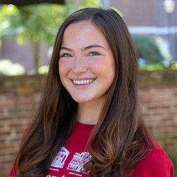 Portrait photo of undergraduate researcher, Lilly Kosoglow, smiling in a garnet t-shirt in front of a historic brick wall on the University of South Carolina Horseshoe.
