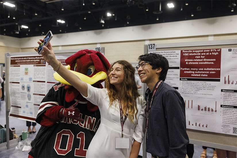 Students pose for a selfie with Cocky in front of their poster presentation at Discover USC 2024
