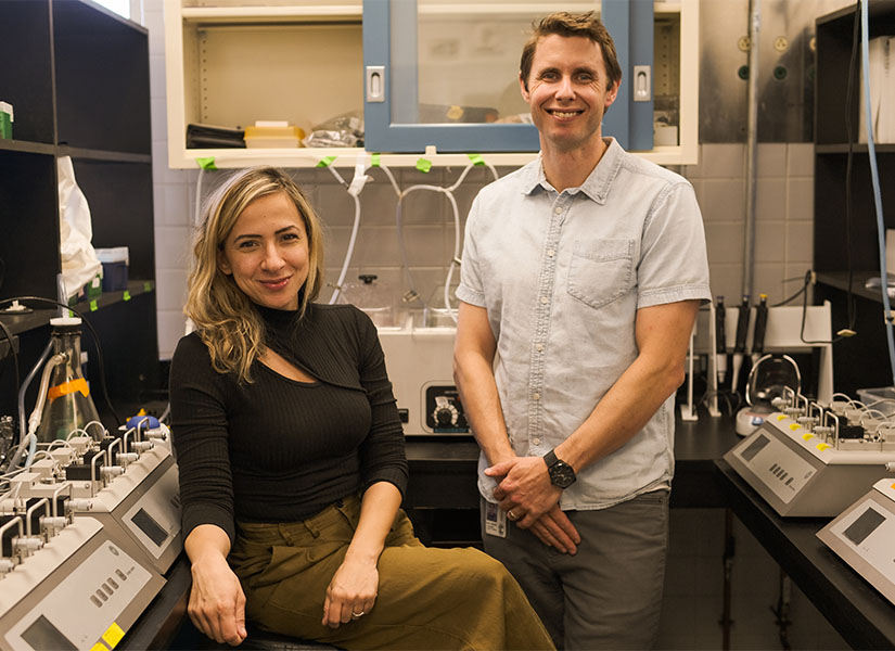 USC cardiovascular researchers Camilla Wenceslau (left) and Cameron McCarthy (right) pose, smiling, for a photo in their laboratory. They are flanked on both sides by research equipment on black lab tables.