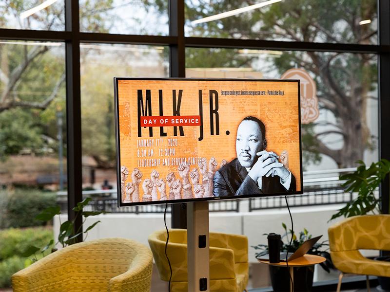 A digital screen displays a Martin Luther King Jr. Day of Service graphic with event details in front of a window facing trees, a ramp and the street.