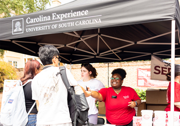 Two Carolina Experience workers talk to students under a black Carolina Experience tent on Greene Street.